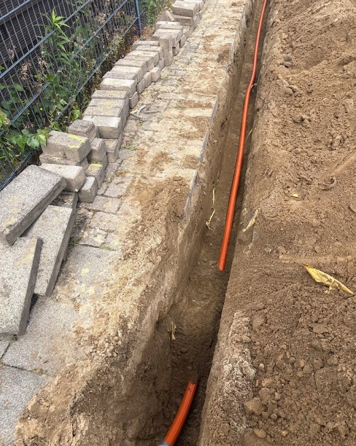 Glasfaser-Leerrohr im offenen Graben entlang eines Gehwegs, aufgenommenes Pflaster daneben